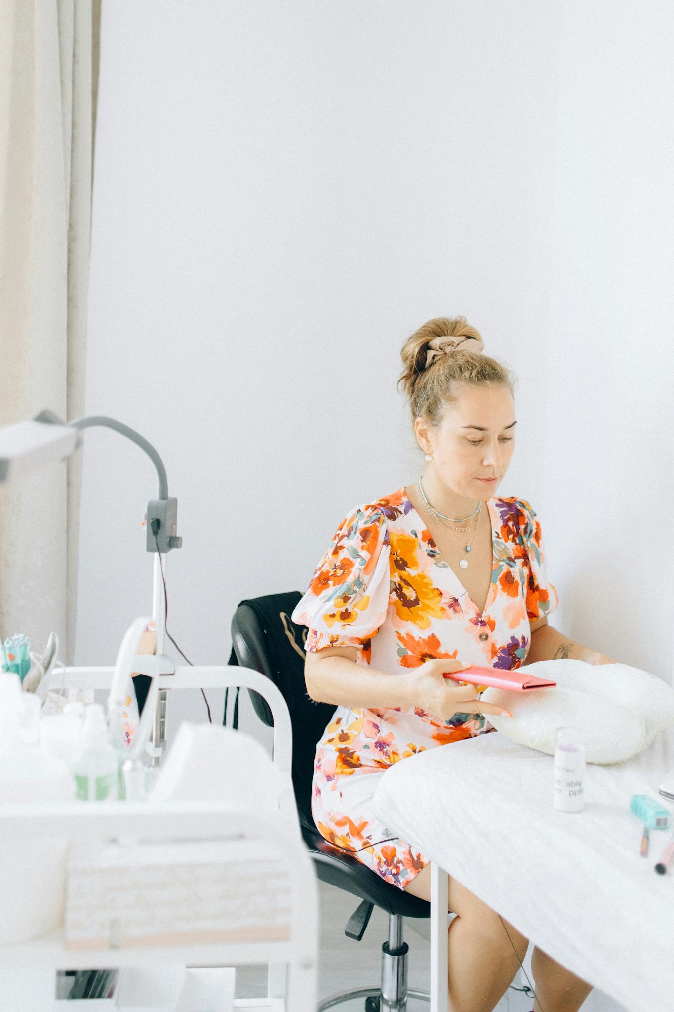 A beautician in a floral dress prepares for eyelash extension treatment in a bright salon.